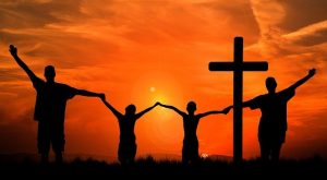 Ein Kreuz und Menschen, die sich an den Händen halten als schwarze Silhouetten vor einem Sonnenuntergang