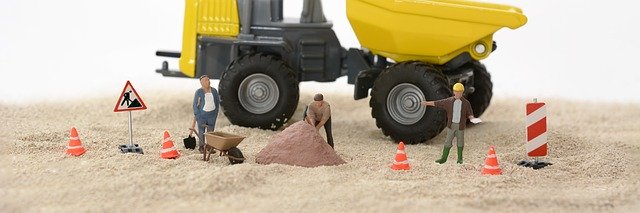 Spielzeugfiguren, -verkehrszeichen und Maschinen sind auf einer Sandfläche als Baustelle drapiert.