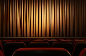 Ein Kinosaal aus der Sicht eines Besuchers. Man sieht Teile der roten Stuhllehnen vor sich und den Vorhang vor der Leinwand, der mit Licht angestrahlt wird.