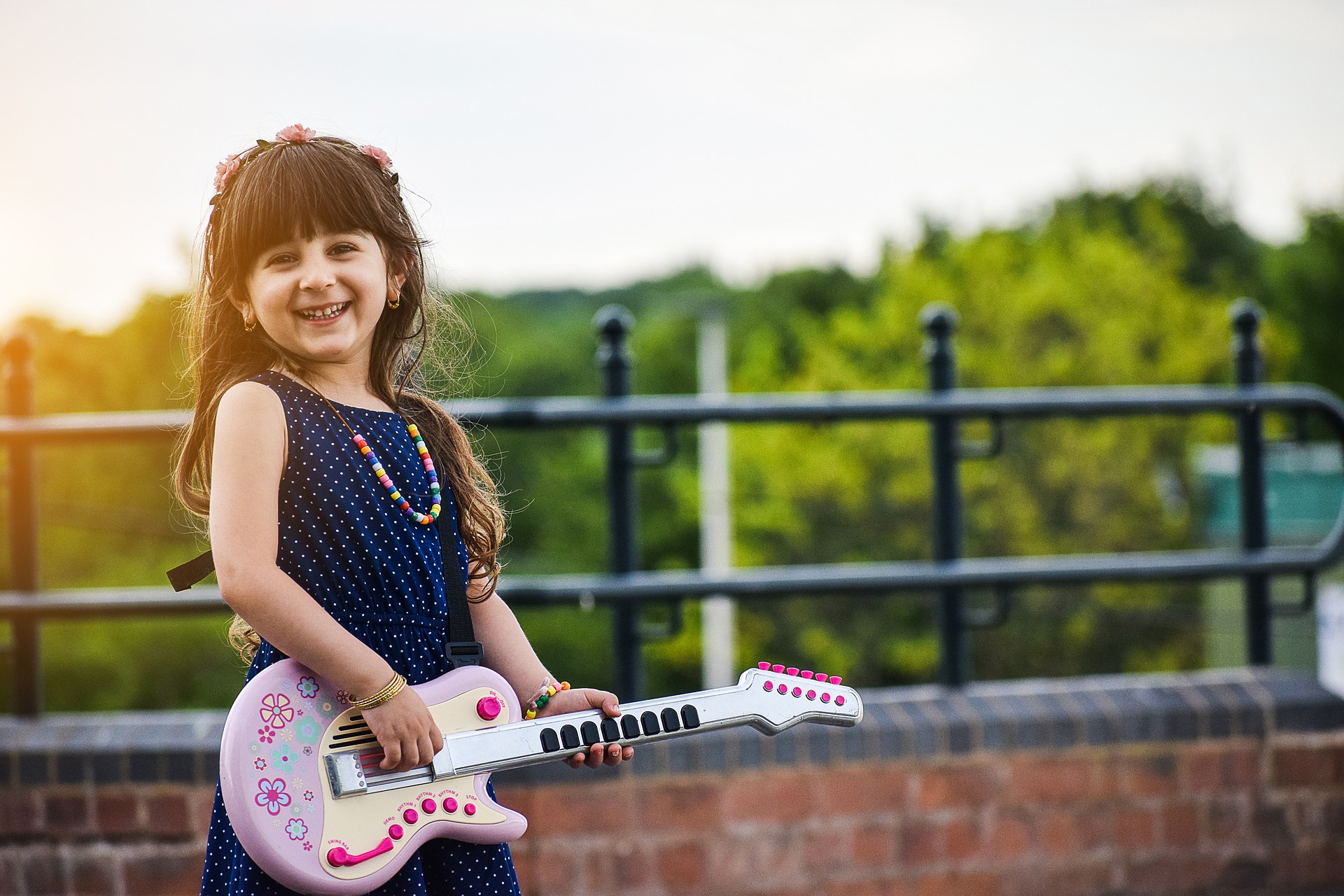 Ein glücklich lächelndes Mädchen steht mit einer rosanen Spielzeuggitarre vor einem Geländer. Im Hintergrund sind Bäume zu sehen. Das Mädchen hat lange Haare, trägt einen Blumenkranz im Haar und eine Kette aus Holzperlen um den Hals. Sie trägt ein dunkelblaues Kleid mit weißen Pünktchen.