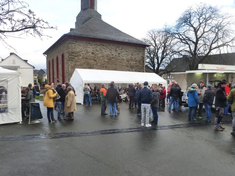 Das Bild zeigt den Manderbacher Weihnachtsmarkt aus dem Jahr 2019. Unter verschiedenen Zelten halten sich Menschen auf und reden miteinander. Im Hintergrund ist die evangelische Kirche zu sehen.