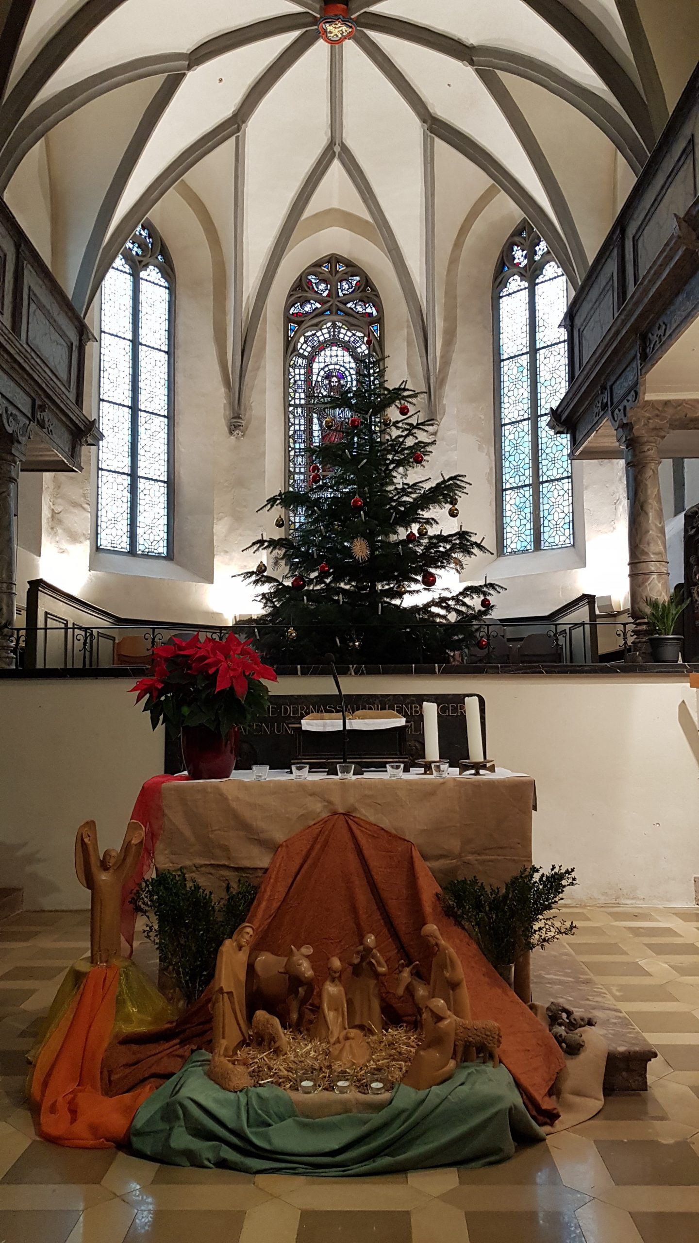 Das Foto zeigt einen Teilbereicht der ev. Stadtkirche Dillenburg mit dem eindrucksvollen Deckengewölbe und den Buntglasfenstern. Vor dem mittleren ist der geschmückte Weihnachtsbaum zu sehen. Weitere im Vordergrund steht der Altar auf dem die Bibel, ein Weihnachtsstern in einem Topf, sowie zwei Kerzen und fünf kleine Gläser mit Teelichtern liegen. Vor dem Altar findet sich die Krippe. Die Figuren sind aus Holz und die Krippenszene ist dargestellt.