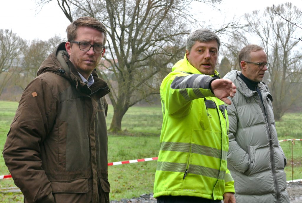 Drei Personen, von denen eine in hellgelber Jacke Ausführungen zum Projekt gibt.