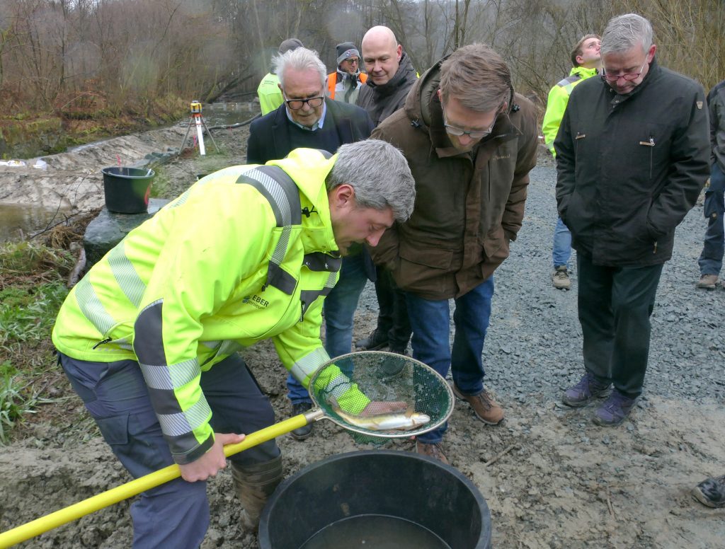Im Vordergrund zeigt ein Mann mit Kescher den weiteren Anwesenden einen Fisch.