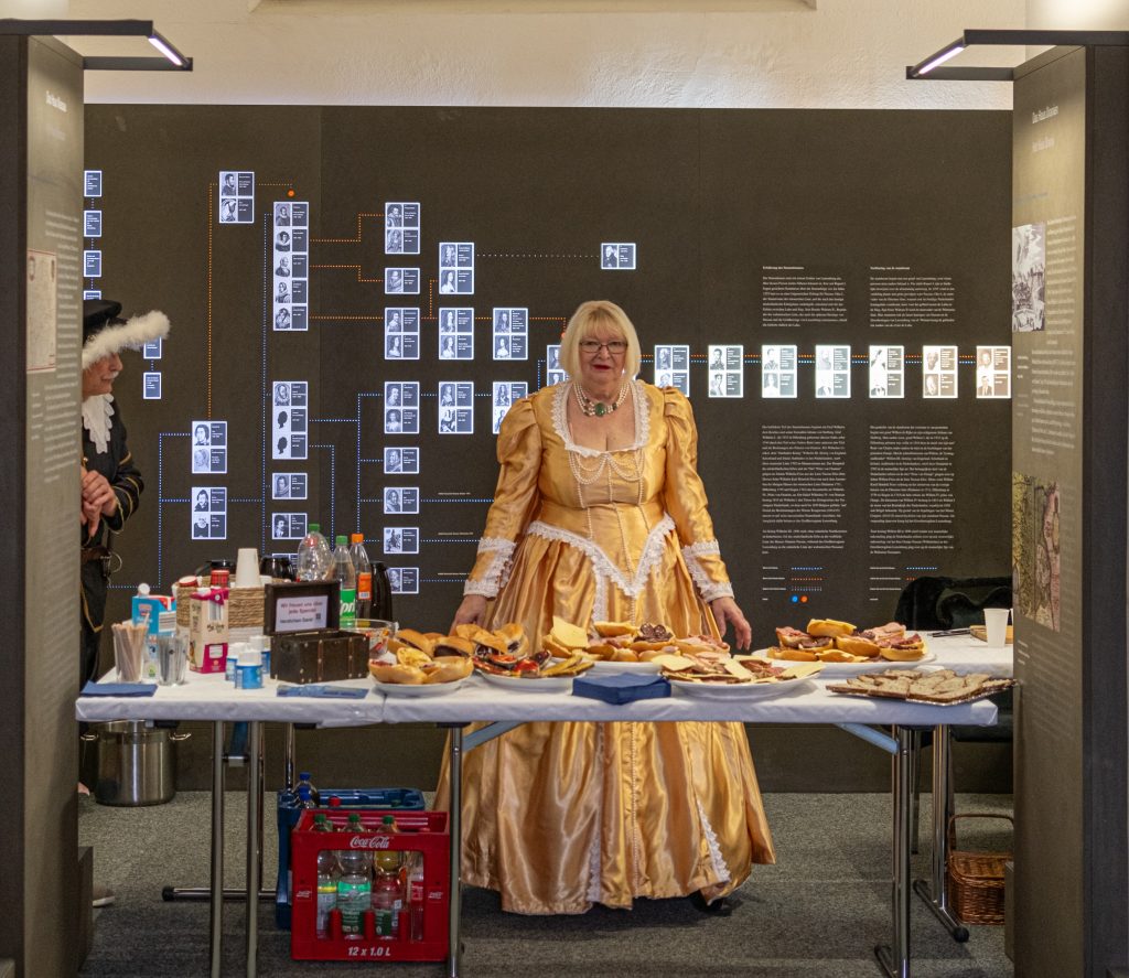 Eine historisch gekleidete Frau im Erdgeschoss des Wilhelmsturms hinter einem Tisch mit Snacks und Getränken.